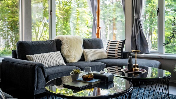 A modern living room with a dark grey sofa, a glass coffee table, and a view of greenery outside. - GROUNDBLOOM YIG STAY (Furano)