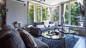 A modern living room with a sofa, two coffee tables, and large windows offering a view of trees. - GROUNDBLOOM YIG STAY (Furano)