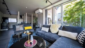 A modern living room with a grey sofa, a coffee table, and a large window offering a view of greenery outside. - GROUNDBLOOM YIG STAY (Furano)