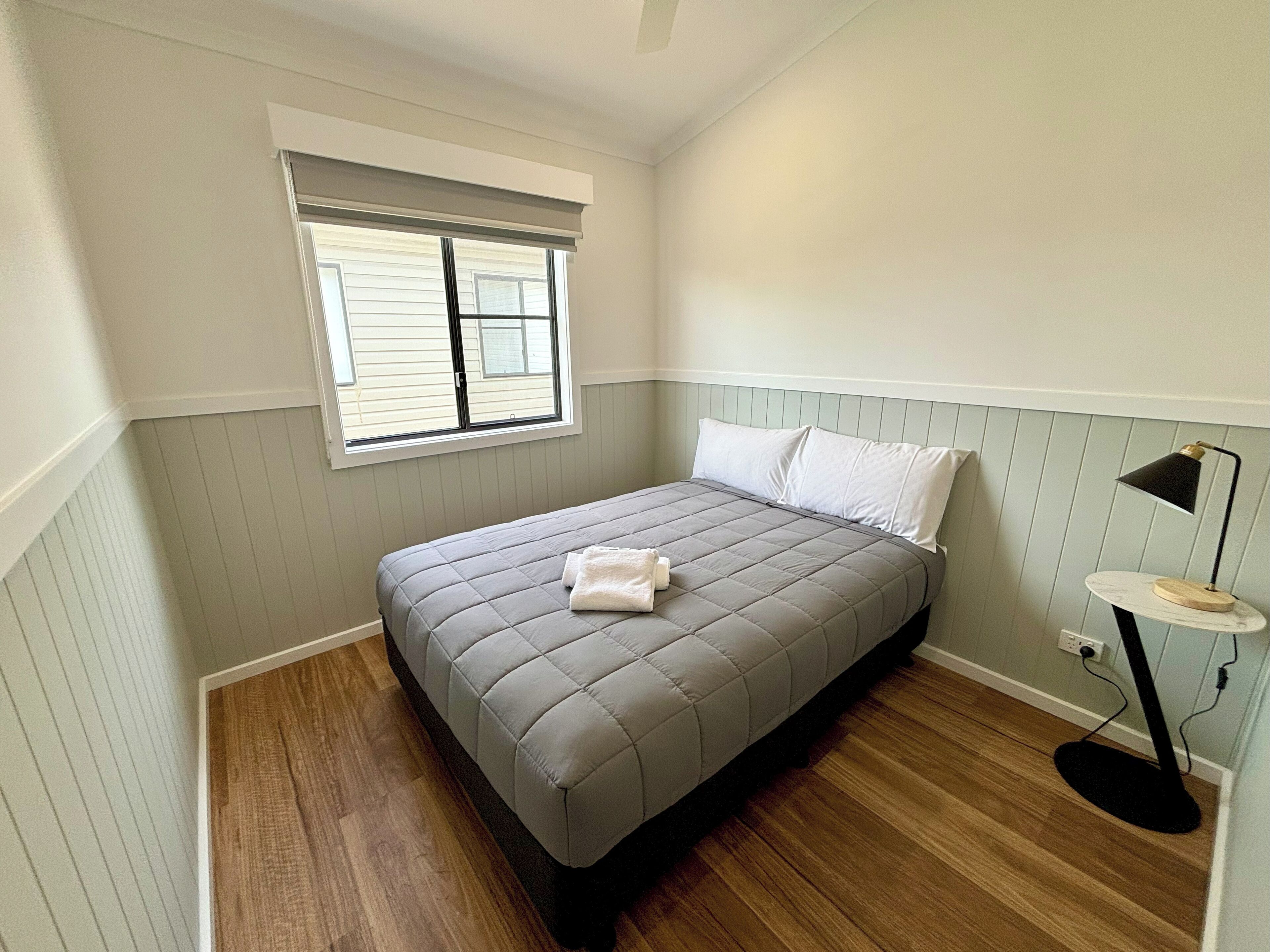A bedroom with a bed, a window, a bedside table with a lamp, and wooden flooring.