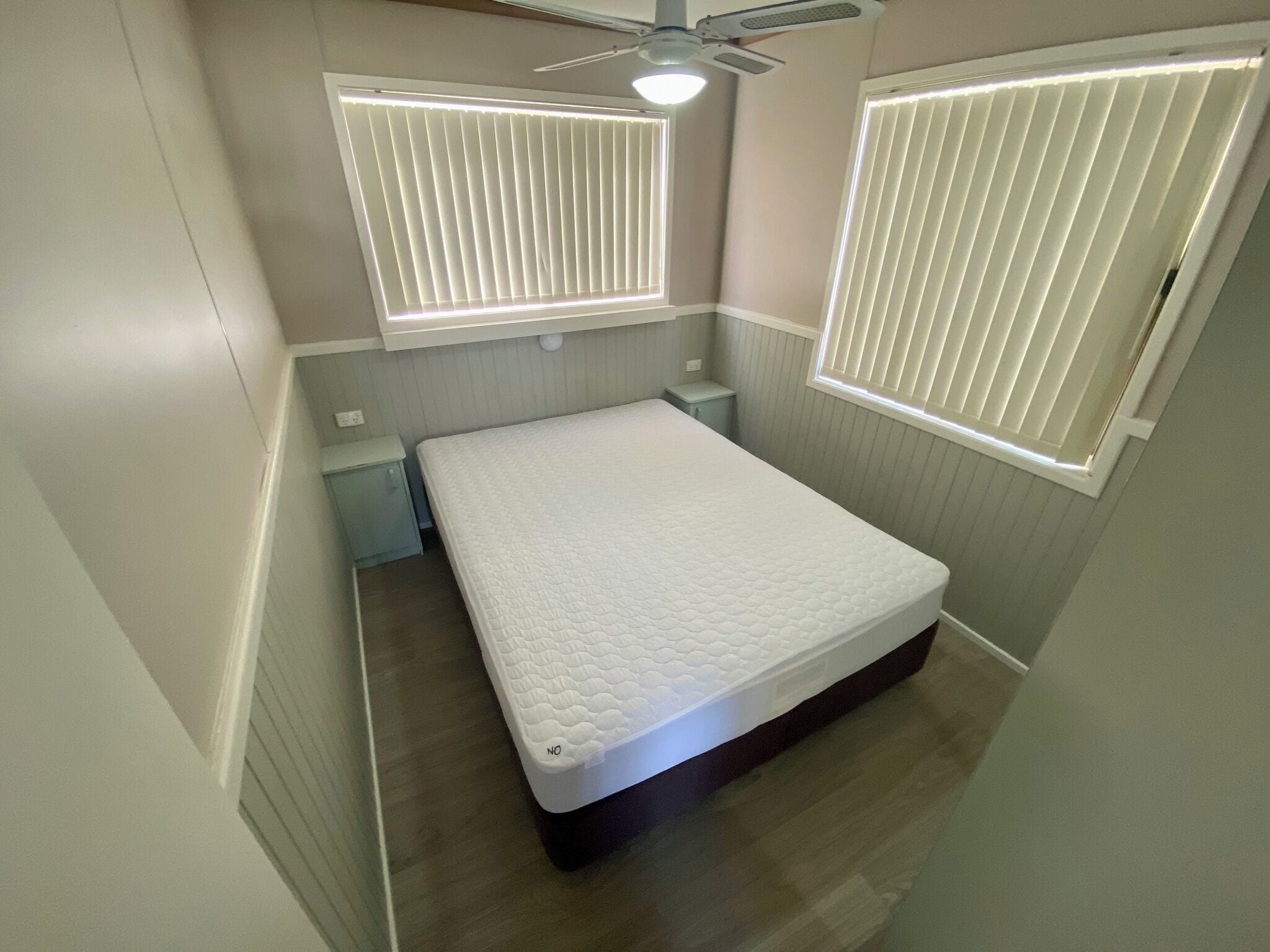 A small, neatly arranged bedroom with a single bed, two bedside tables, and two windows with blinds.