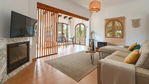 Un salon avec une cheminée, un canapé et un téléviseur. Une porte en bois donne accès à un espace extérieur aménagé avec une table et des chaises.