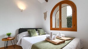 Un lit bien fait, avec une literie verte et blanche, une table de chevet ornée d’une plante et d’un livre, et une fenêtre donnant sur un mur de pierre et de la verdure.