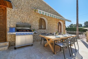 Une terrasse extérieure avec un mur en pierre, un barbecue, une table en bois dressée pour le repas et des chaises en métal.
