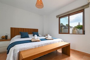 Une chambre à coucher avec un lit en bois, deux oreillers, un banc, une table de chevet et une fenêtre donnant sur l’extérieur.
