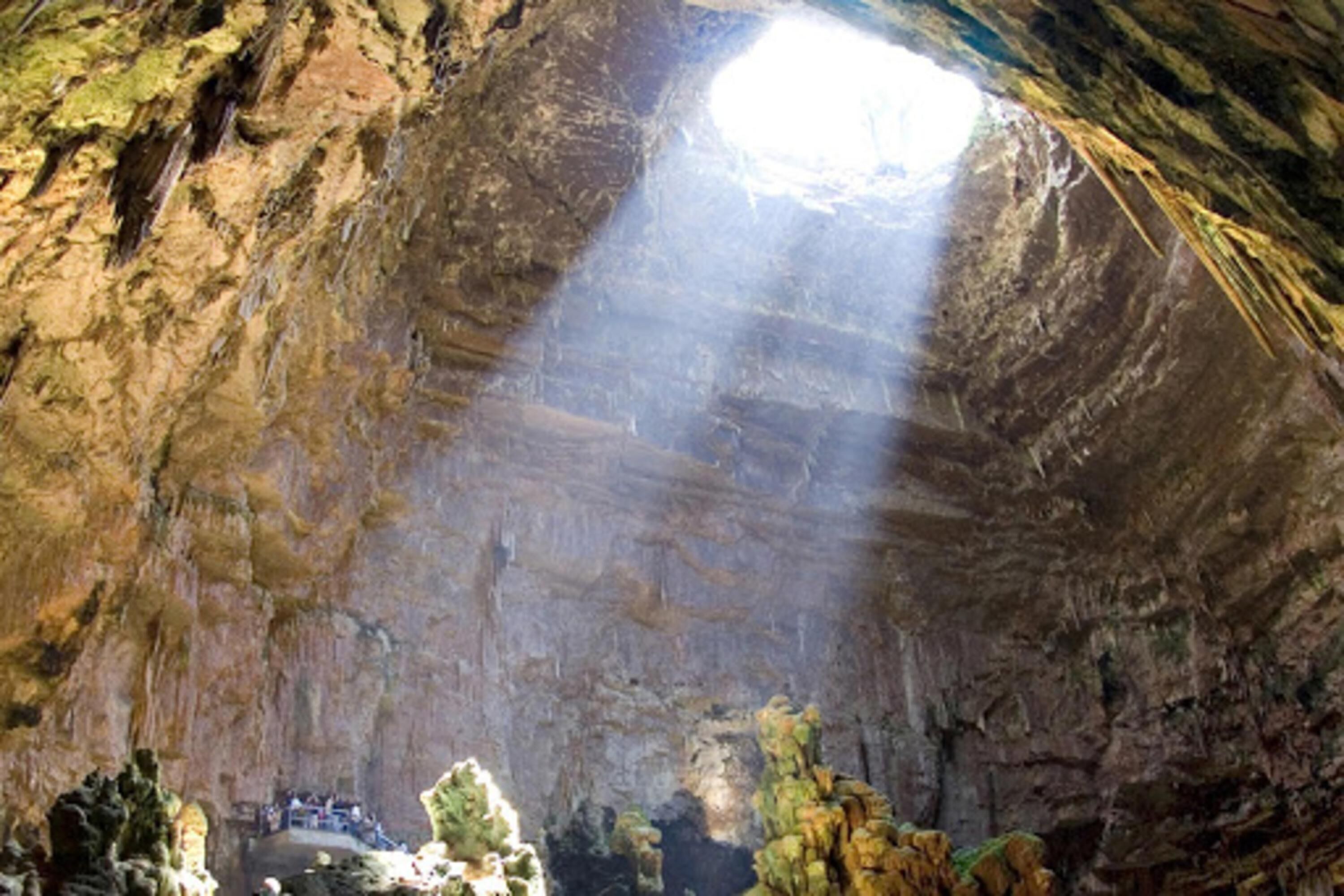 A cave with stalactites and stalagmites, a small platform with people, and a natural light source from above.