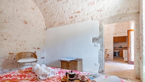 A room with a bed, a wicker chair, a small table with a tray of towels and a coffee pot, and a view of a kitchen through an open doorway. - Lamia Terra 3br, Terrace & BBQ (Castellana Grotte)