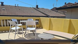 A sunlit outdoor patio with a round table and white chairs, a yellow wall, and a tiled roof in the background.