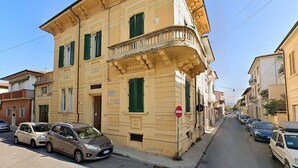 A narrow street lined with parked cars and buildings with balconies and green shutters.