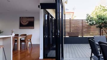 An interior space with a wooden dining table and chairs, a white countertop, and a glass door leading to an outdoor deck with a wooden fence and greenery.
