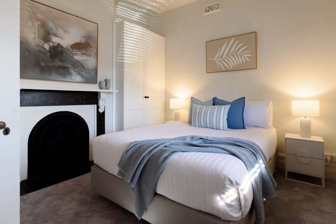 A bedroom with a bed, bedside lamps, a fireplace, and artwork on the walls.