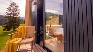 A wooden deck with a wicker chair and a yellow throw blanket, a glass door leading to a bathroom with a bathtub and a bottle of champagne, and a view of a forested landscape. - The Valley Resort (Vama Buzăului)
