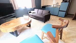 A modern living room with a wooden dining table, blue chairs, a grey sofa, and a kitchen area with blue cabinets. - Izu Nagaoka The NooK - Family Stay with Hot Spring, Work-friendly & Long Stay (Izunokuni)