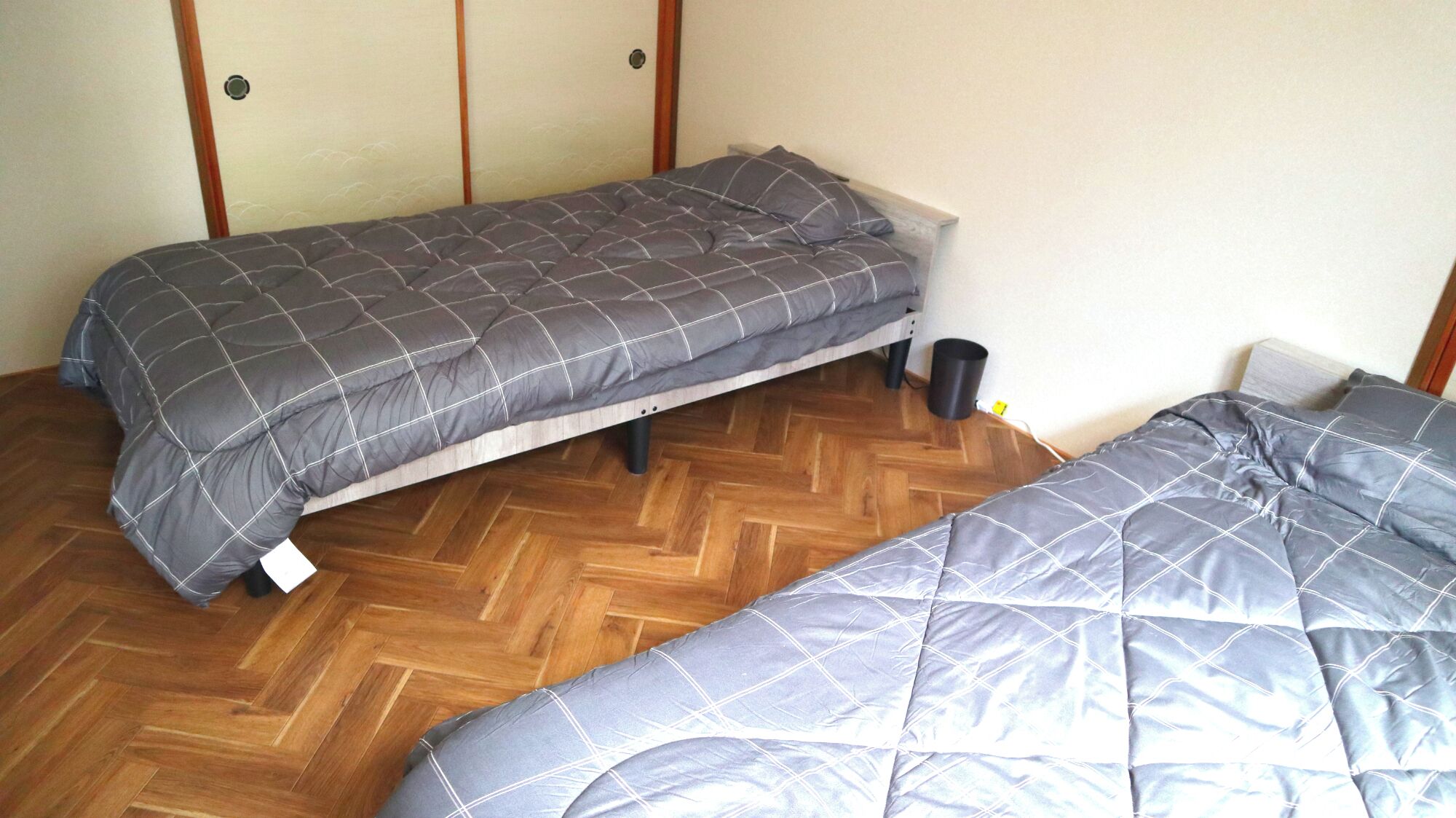 Quarto pequeno com duas camas de solteiro, piso de madeira e parede lisa.