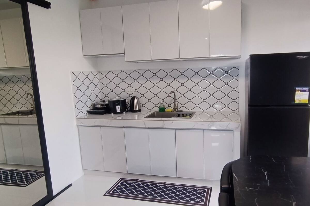 A compact kitchen with white cabinets, a black refrigerator, a sink, and a countertop with a coffee maker and kettle.