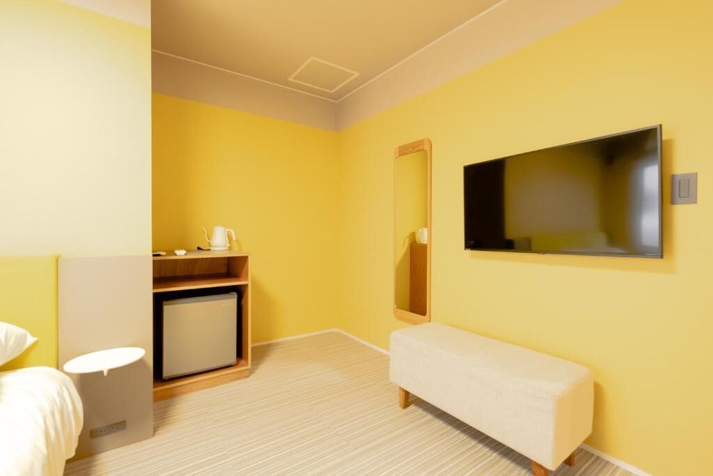 A hotel room with a yellow wall, a flat-screen TV mounted on the wall, a small wooden cabinet, a white bench, and a white bedside table.