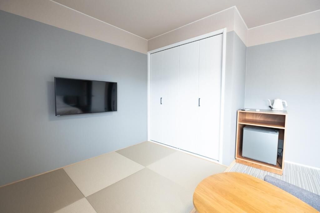 A modern room with a flat-screen TV mounted on the wall, a sliding wardrobe, a small wooden table with a kettle, and tatami flooring.