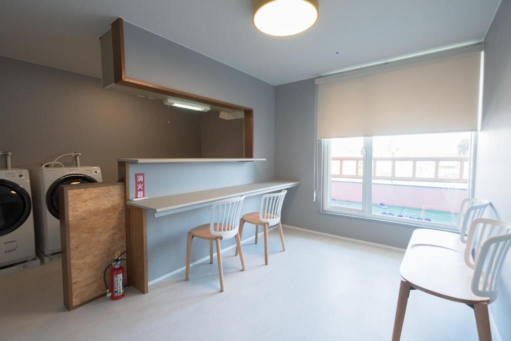 A laundry room with a washing machine, dryer, and fire extinguisher. There are two chairs and a table with a window in the background.