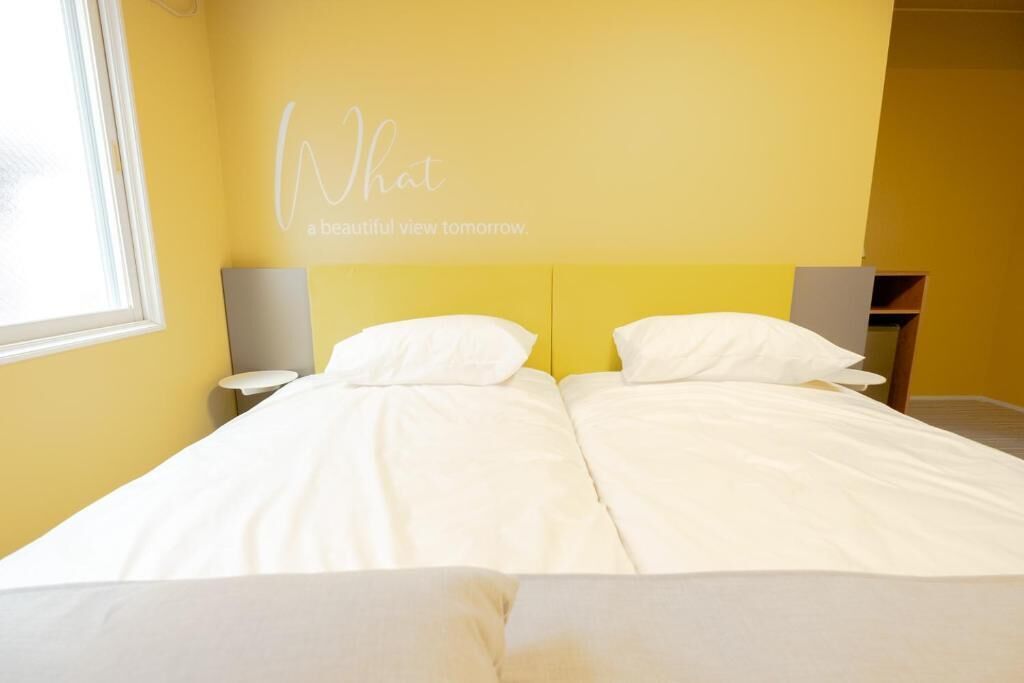 Una cama doble con ropa blanca en una habitación con paredes amarillas y una ventana.