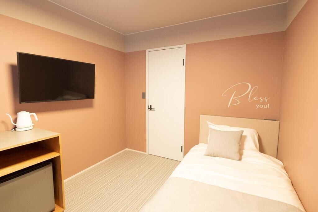 A hotel room with a bed, a TV mounted on the wall, a bedside table with a kettle, and a door with a "Bless you!" sign.