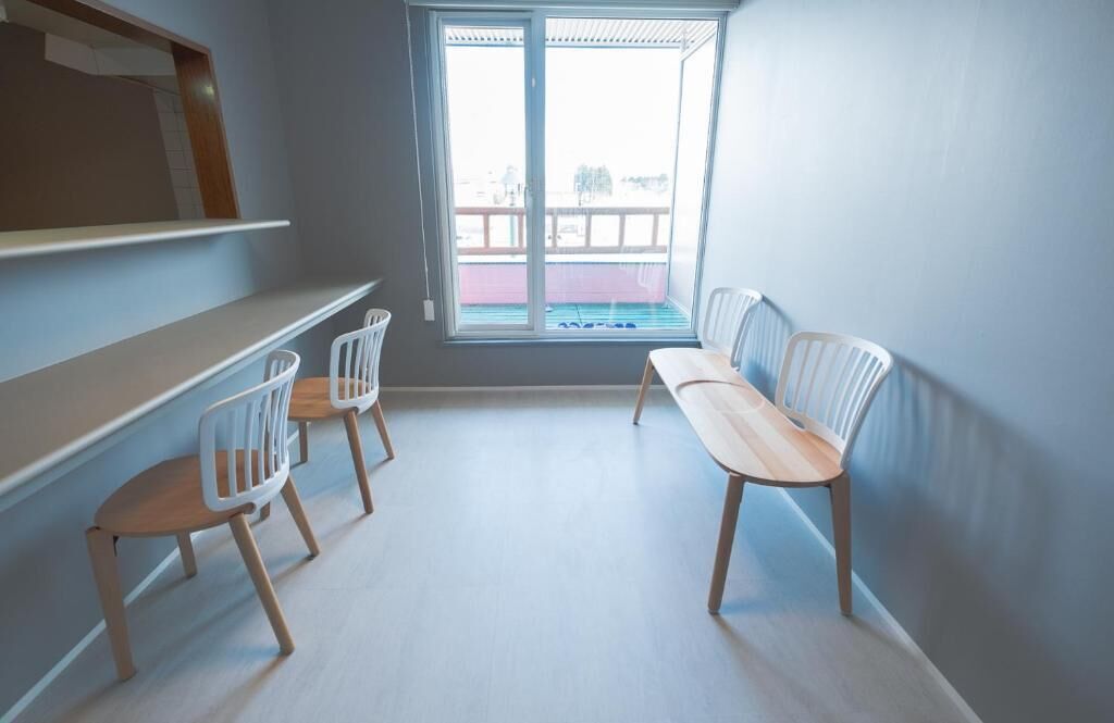 A room with a large window, a wooden bench, and two white chairs.
