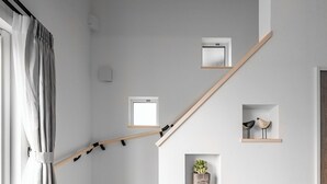 Una escalera de madera con barandilla, bordeada por paredes blancas y una ventana con cortinas.