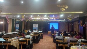 A conference room with tables and chairs, a stage with a presenter, and a screen displaying a presentation. - Green Castle (Panipat)