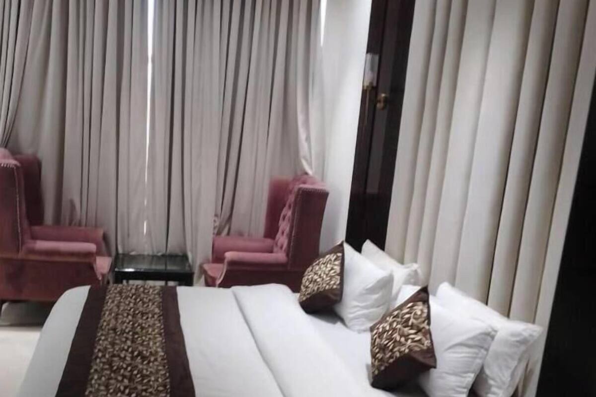 A modern hotel room with a large bed, two armchairs, a small table, and a large window with curtains.