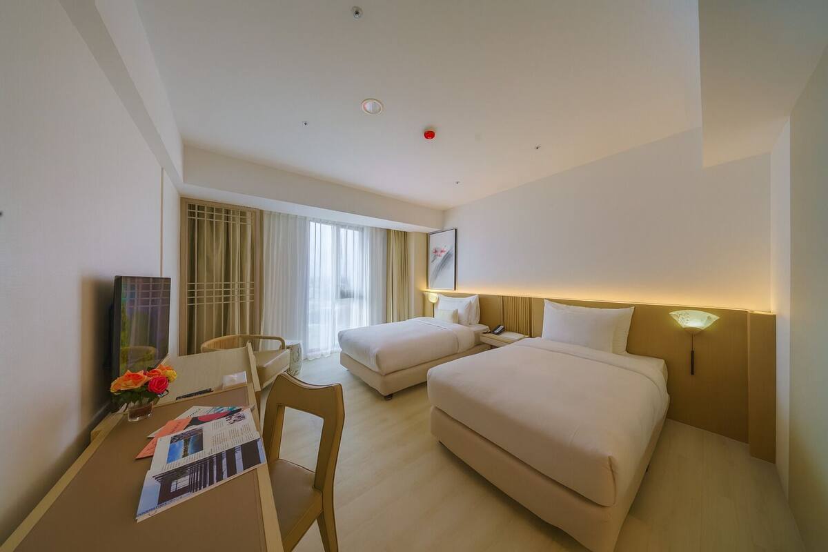 A hotel room with two beds, a desk with a chair, a small table with a vase of flowers, and a large window with curtains.