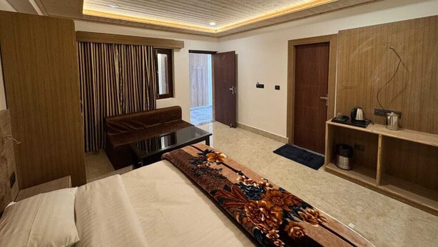 A modern bedroom with a bed, a bench, a wooden wardrobe, and a door leading to a balcony. - Hotel Kuber Palace Badrinath (Badrinath)