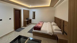 A bedroom with a wooden ceiling, a bed with a red blanket, a bedside table, and a small desk with a chair. - Hotel Kuber Palace Badrinath (Badrinath)
