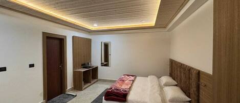 A bedroom with a wooden ceiling, a bed with a red blanket, a bedside table, and a small desk with a chair.