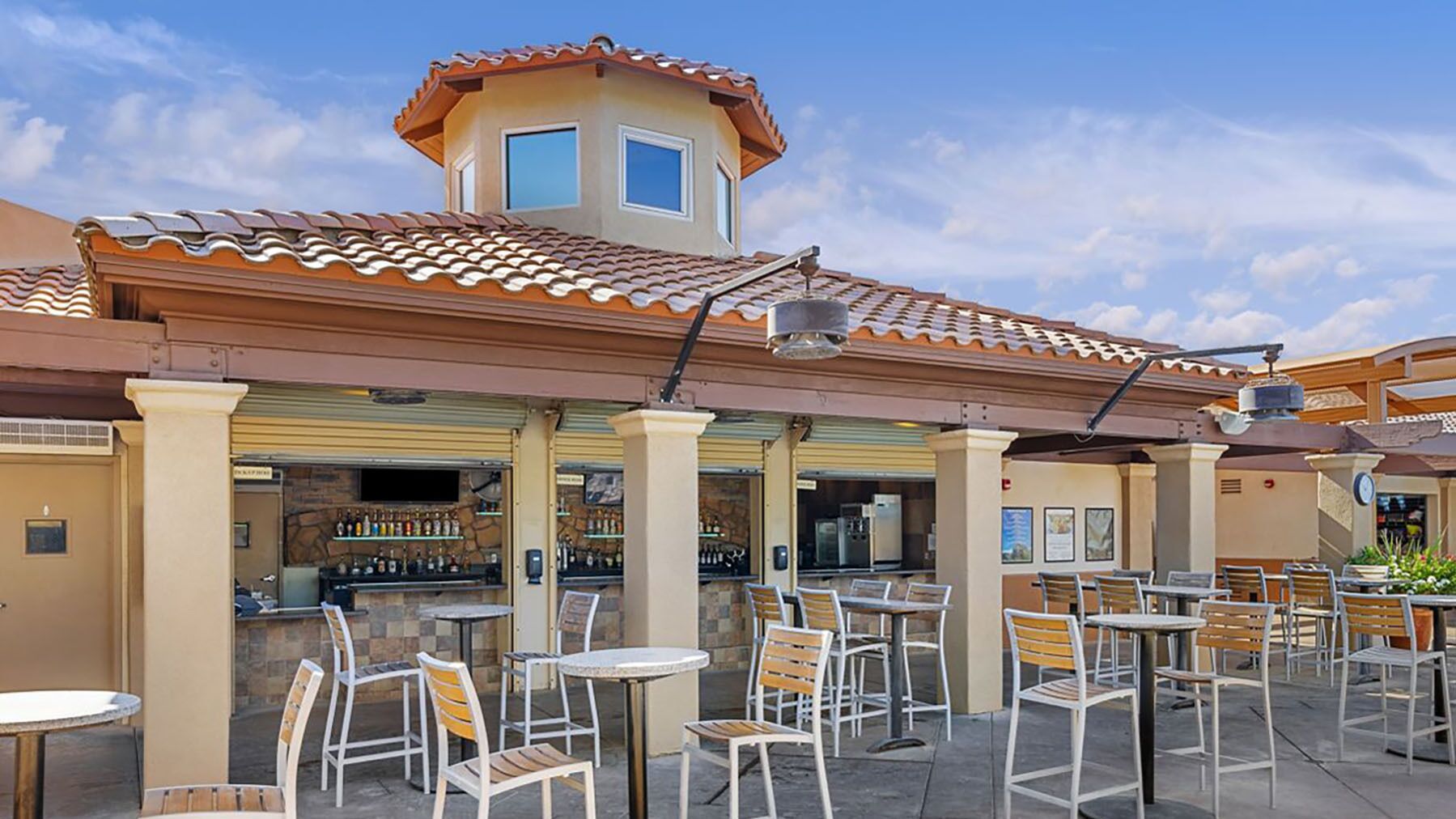 Un espace extérieur aménagé en terrasse, avec des tables et des chaises, un bar doté d’étagères remplies de bouteilles, et un bâtiment avec un toit en tuiles et une petite structure ressemblant à une tour.