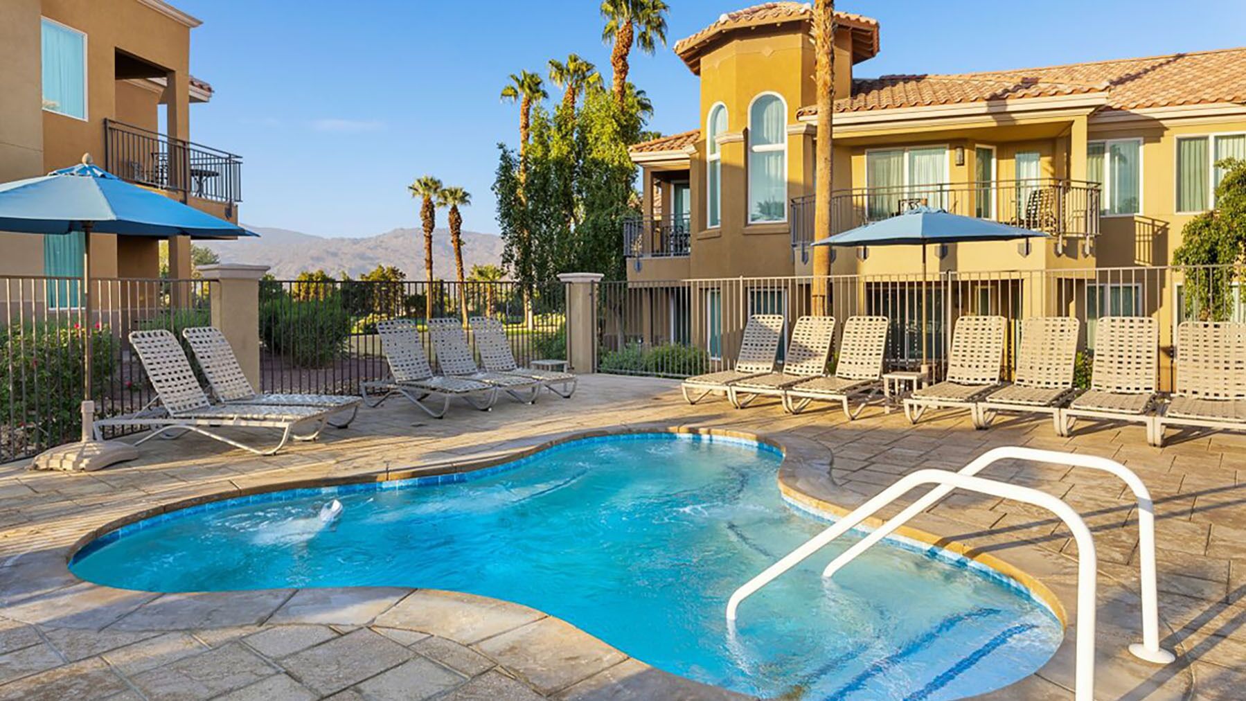 A swimming pool with lounge chairs and umbrellas, surrounded by a building with balconies and palm trees.