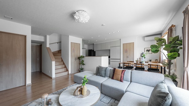 A modern living room with a grey sectional sofa, a round coffee table, and a staircase leading to an upper floor. - THE VERTEX (Asahikawa)