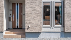 A modern building entrance with a stone facade, a wooden door, and a window with a view of the interior. - THE VERTEX (Asahikawa)