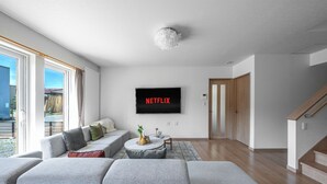 A modern living room with a grey sofa, a coffee table, and a flat-screen TV mounted on the wall displaying "NETFLIX". - THE VERTEX (Asahikawa)
