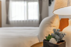 A neatly made bed with a white comforter and pillows, a bedside table with a potted succulent, and a lamp with a beige lampshade. - THE VERTEX (Asahikawa)