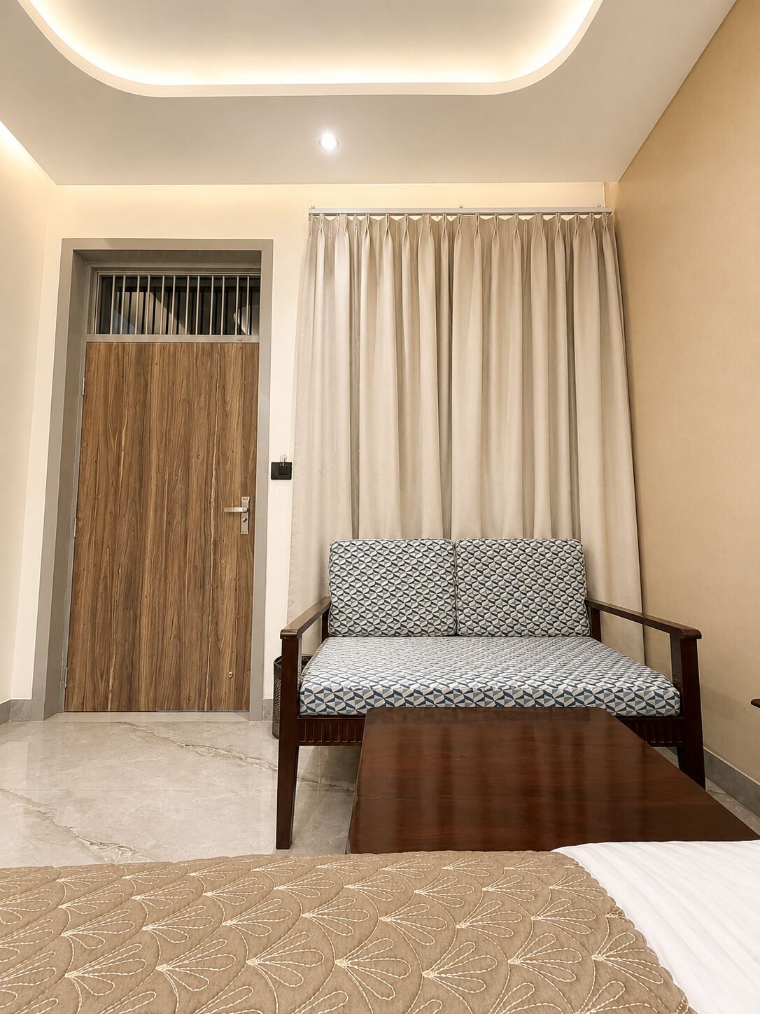 A room with a wooden door, a sofa with patterned cushions, a wooden coffee table, and a bed with a patterned bedspread.