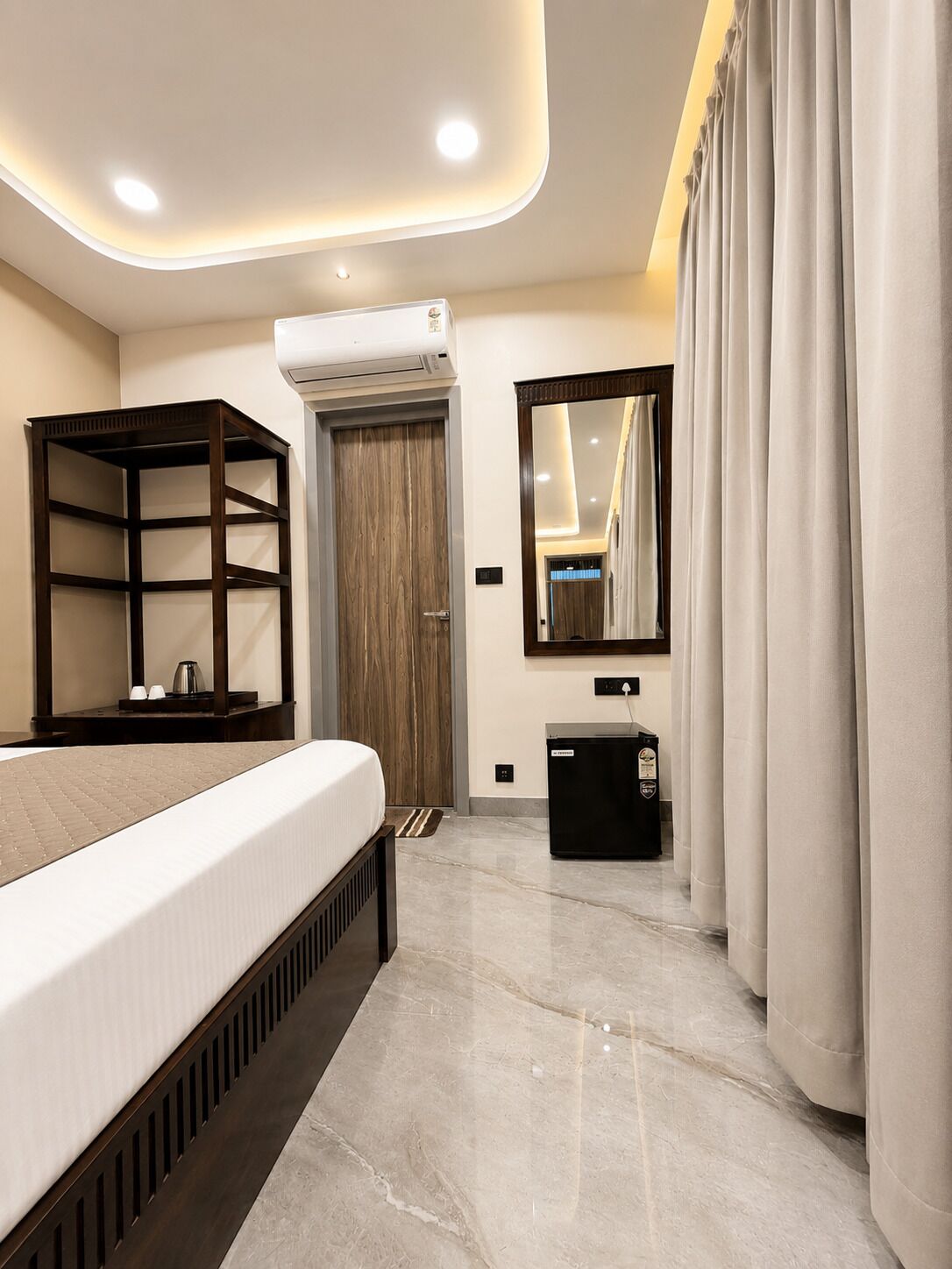 A modern hotel room with a bed, built-in shelving, a small desk, a mirror, and an air conditioning unit.