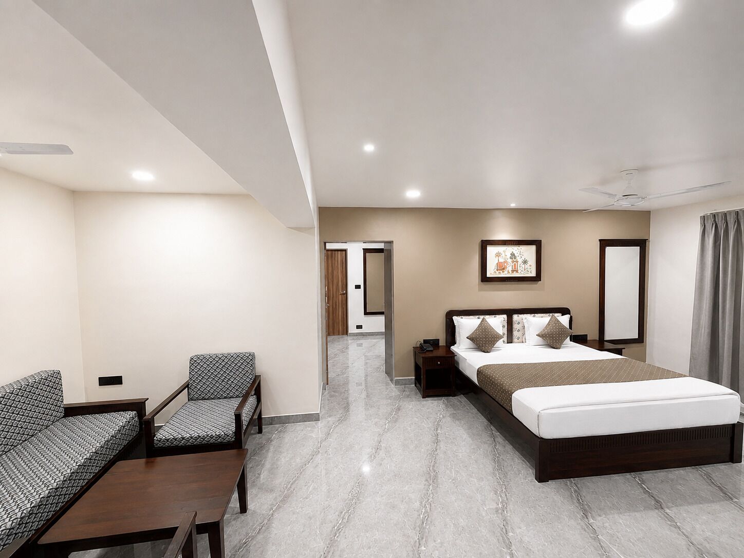Habitación de hotel con una cama, un escritorio, dos sillas, una mesita de noche, un ventilador de techo y un cuadro en la pared.