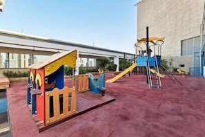 A playground with a slide and climbing structures, located on a rooftop with a view of buildings in the background.