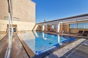 A rooftop swimming pool with clear blue water, surrounded by lounge chairs and a modern building with large windows.