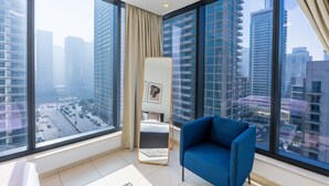 Habitación de hotel moderna con grandes ventanales que ofrecen vistas a la ciudad, un sillón azul, una mesita auxiliar y un cuadro en la pared.