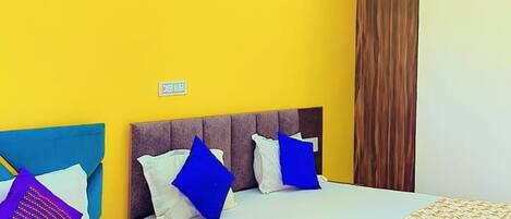 A hotel room with two beds, each with a wooden headboard, white bedding, and blue and purple pillows. The room has a yellow accent wall and a ceiling fan.