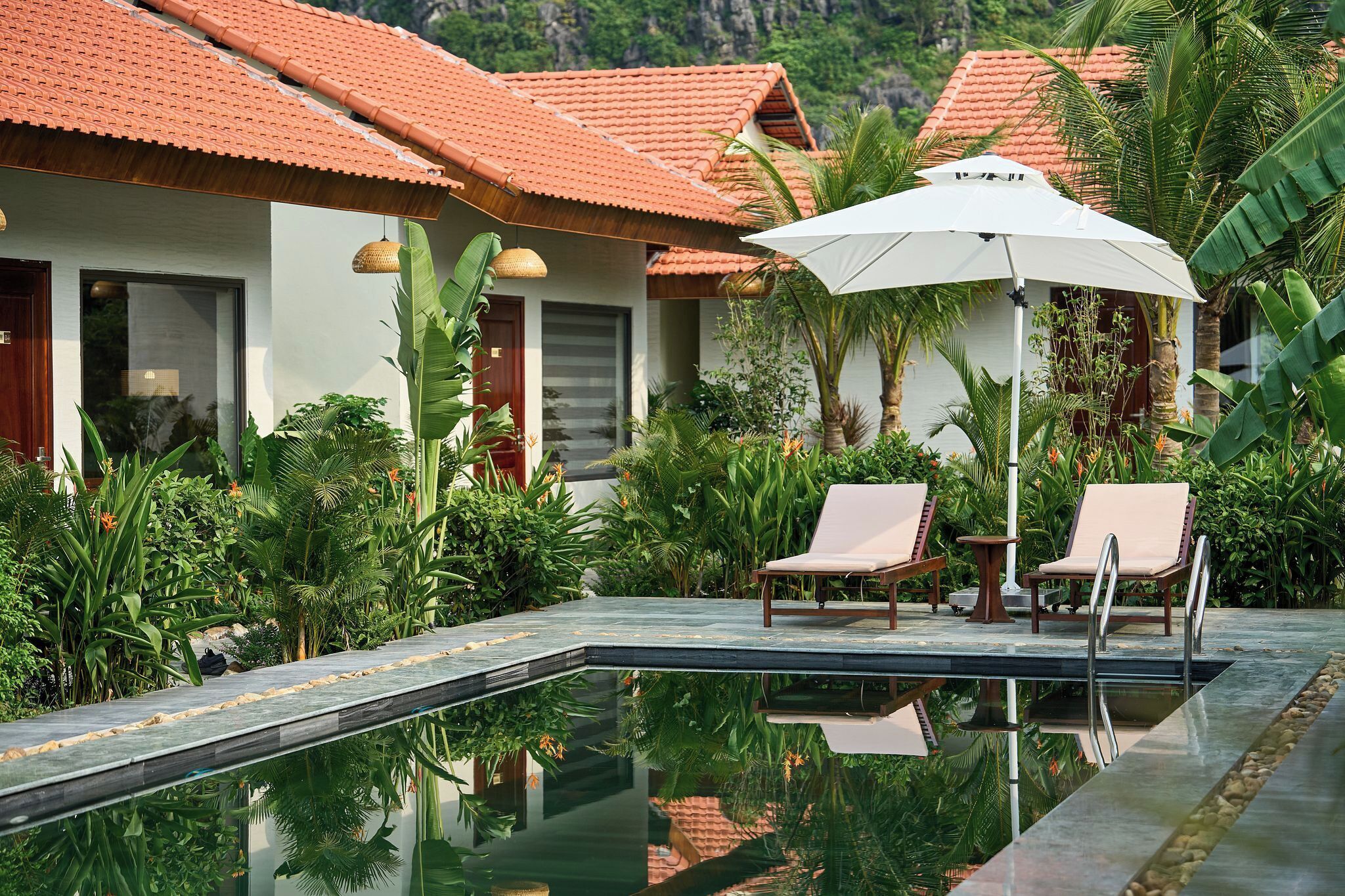 Un espace aménagé au bord de la piscine, avec deux chaises longues et un parasol blanc, entouré d’une végétation tropicale luxuriante et d’une maison dont le toit est recouvert de tuiles rouges.