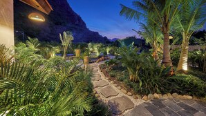 A well-lit garden with a stone pathway, palm trees, and a mountain backdrop. - The Greenary Garden (Ninh Binh)