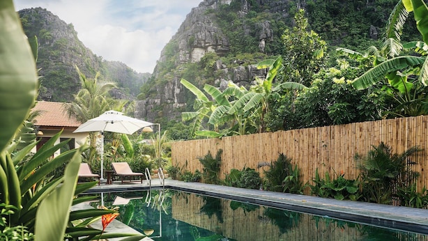 A swimming pool with lounge chairs and an umbrella, surrounded by lush greenery and a bamboo fence. - The Greenary Garden (Ninh Binh)