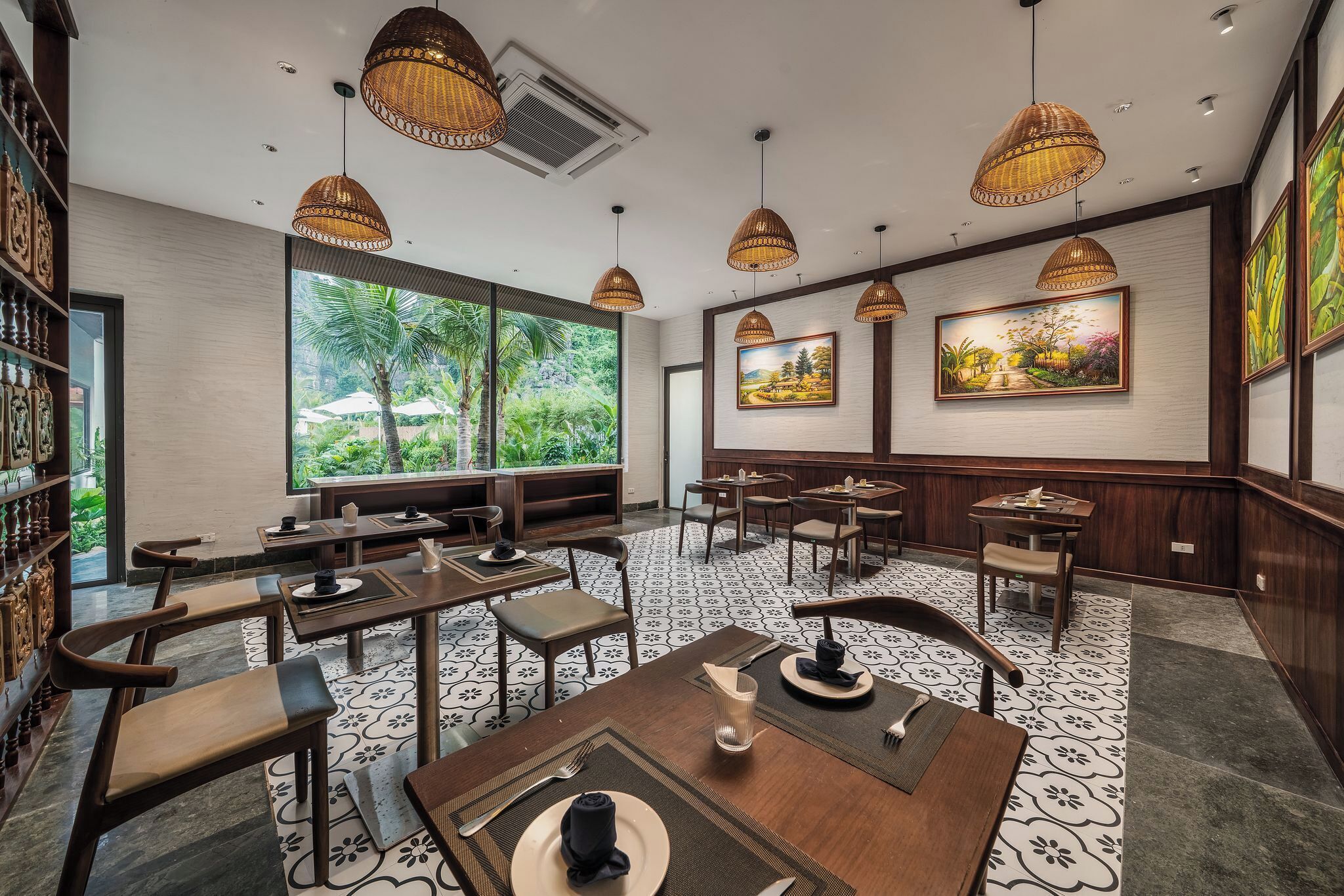 A restaurant with wooden tables and chairs, large windows with a view of greenery, and hanging wicker lamps.