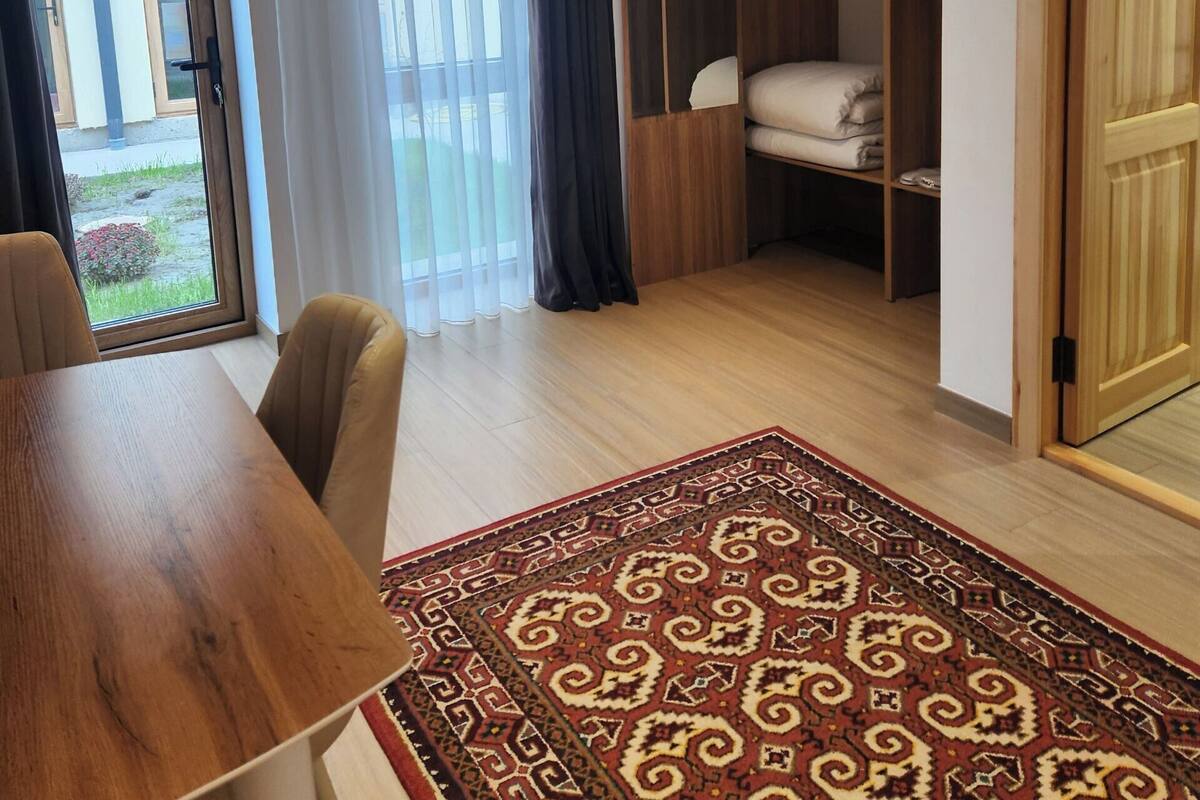 A room with a wooden table, a patterned rug, a bed with a white mattress, a wooden wardrobe, and a sliding glass door leading to an outdoor area.